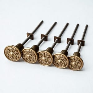 May include: Five antique brass cabinet knobs with ornate floral designs. Each knob has a round face with a detailed pattern, a cylindrical base, and a threaded rod for installation. The knobs are arranged in a row against a white background.