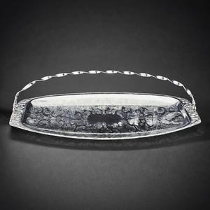 May include: A silver serving tray with an engraved floral pattern. The tray has a curved rectangular shape and a twisted handle. The edges are decorated with detailed patterns. The background is black. The tray is approximately 30 cm long.