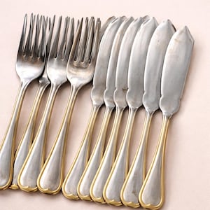 Peut inclure: Un ensemble de couverts argentés composé de trois fourchettes et cinq couteaux. Chaque pièce présente une tête argentée polie et un manche avec une bordure dorée. Les manches ont un design lisse et arrondi avec une petite encoche à la base.
