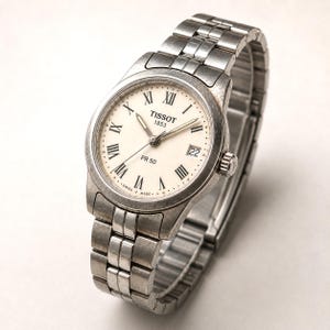 May include: A silver Tissot PR 50 watch with a cream-colored face, Roman numeral hour markers, and a date display at the 3 o'clock position. The watch has a stainless steel band and is set against a white background.