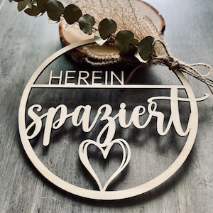 Könnte beinhalten: Runder Holzschild mit dem deutschen Schriftzug "HEREIN spaziert" und einem Herz-Ausschnitt. Das Schild ist aus hellem Holz gefertigt und mit getrocknetem Grün und einer Holzscheibe verziert.