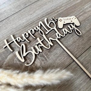 Könnte beinhalten: Ein Kuchenaufsatz aus Holz mit den Worten "Happy 16th Birthday" in Schreibschrift. Ein kleiner Holzcontroller mit dem Namen "Luca" ist angebracht. Der Aufsatz ist hellbraun und hat einen Holzstab zum Einstecken in einen Kuchen.