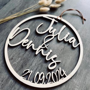 Könnte beinhalten: Ein Holz-Kreis mit den Namen "Julia" und "Dennis" in Kursivschrift, sowie dem Datum "21.09.2024".