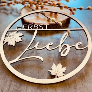 Könnte beinhalten: Ein kreisförmiges Holzschild mit dem Wort HERBST in Blockbuchstaben und dem Wort Liebe in Schreibschrift. Das Design enthält zwei Ahornblatt-Ausschnitte, einen oben links und einen unten rechts, die alle von einem dünnen Holzkreis umrahmt sind.
