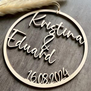Könnte beinhalten: Ein kreisförmiges Schild aus hellem Holz mit den lasergeschnittenen Namen Kristina und Eduard, die durch ein Et-Zeichen in der Mitte verbunden sind. Das Datum 16.08.2024 ist unten ausgeschnitten. Eine kleine Schlaufe oben dient zum Aufhängen.
