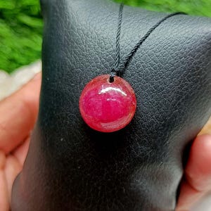 May include: A circular, polished, deep pink gemstone pendant suspended from a thin black cord. The stone features a smooth, reflective surface with subtle internal variations in color intensity, showcasing a vibrant magenta hue throughout the round shape.