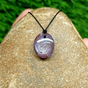 May include: A polished oval-shaped gemstone pendant with a deep reddish-purple hue and a visible star-like light reflection pattern. The stone is suspended from a thin black cord, showcasing a smooth, reflective surface with internal inclusions.
