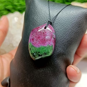 May include: A pendant featuring a polished, irregular-shaped stone with a distinct colour split of vibrant magenta-pink and deep forest green, suspended from a thin black cord. The stone has a smooth, glossy surface texture and a natural appearance.