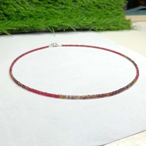 May include: A delicate necklace composed of tiny, faceted beads in a gradient of colours, transitioning from vibrant pink and red tones to earthy brown and amber hues. The strand is circular and features a small, silver-toned metal clasp closure.