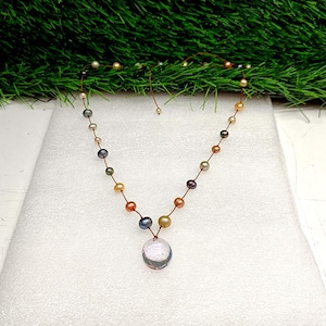 Op de afbeelding: Een delicate ketting met een streng kleurrijke parels in tinten roze, groen en goud. De ketting heeft een heldere, ronde hanger in het midden. De ketting wordt gepresenteerd op een wit oppervlak met groen gras op de achtergrond.