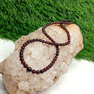 Pode incluir: Um conjunto de colar e pulseira com pequenas contas redondas de cor vermelho escuro. As contas são enfiadas juntas, formando um conjunto de joias delicado e elegante. As joias são exibidas em uma pedra áspera de cor clara sobre um fundo verde.