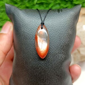 Op de afbeelding: Een marquisevormige zonnesteen hanger met een doorschijnend lichaam en levendige oranje en roodbruine metalen insluitingen. De edelsteen is glad gepolijst en hangt aan een dun zwart koord, wat natuurlijke lichtreflecties laat zien.