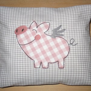 Könnte beinhalten: Ein graues und weißes kariertes Kissen mit einem rosa und weißen karierten Schwein, das darauf gestickt ist. Das Schwein hat einen grauen Flügel und einen eingerollten Schwanz.