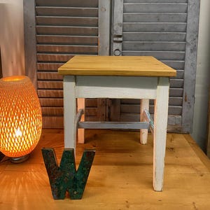 Könnte beinhalten: Ein kleiner, quadratischer Holzschemel mit einer hellen Oberseite und weißen Beinen. Die Beine sind mit grauer Farbe akzentuiert. Der Hocker ist etwa 46 cm hoch. Der Hintergrund zeigt graue Holzläden und eine dekorative Lampe.