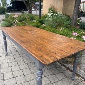 Könnte beinhalten: Ein rechteckiger Tisch mit einer natürlichen, verwitterten braunen Holzplatte mit sichtbarer Maserung und Textur. Der Tisch wird von vier gedrechselten, spindelförmigen Beinen getragen, die in einem matten Grau lackiert sind und einen Kontrast bilden.