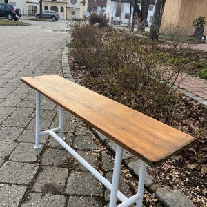 Könnte beinhalten: Eine lange Holzbank mit hellbrauner Oberfläche und weißen Metallbeinen. Die Bank befindet sich im Freien auf einer Ziegel- und Steinoberfläche, mit einem Hintergrund aus Gebäuden und Bäumen.