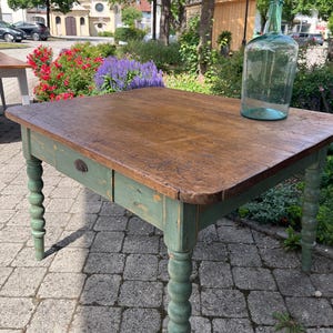 Könnte beinhalten: Ein quadratischer Holztisch mit einer naturfarbenen braunen Platte und einem salbeigrünen Untergestell, das vier gedrechselte Beine und eine einzelne Schublade mit einem kleinen Metallgriff aufweist. Eine große, klare, hellblaue Glasflasche steht darauf.