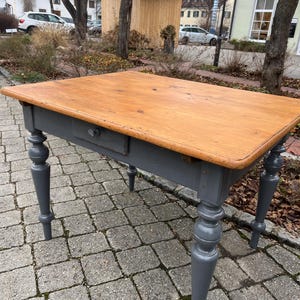 Könnte beinhalten: Ein rechteckiger Holztisch mit einer natürlichen Holzplatte und einem grau lackierten Untergestell. Der Tisch hat eine einzelne Schublade und gedrechselte Beine. Der Tisch ist ca. 152 cm lang, 76 cm breit und 76 cm hoch.