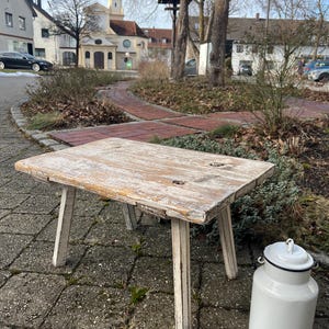 Könnte beinhalten: Ein weiß gestrichener Holzhocker mit vier Beinen und einer rechteckigen Oberseite. Der Hocker steht auf einem Ziegelhof vor einem Gebäude.