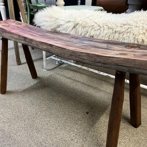 Könnte beinhalten: Eine rustikale Holzbank mit einer geschwungenen Oberseite und einer natürlichen Holzoptik. Die Bank hat auf jeder Seite drei Beine, die aus dunklem Holz gefertigt sind. Die Bank steht auf einem Teppichboden.