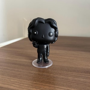 May include: A matte black vinyl collectible figure featuring a stylized head with wavy hair, a small triangular nose, and two small white circular eyes. The figure wears a simple shirt with a chest pocket and pants, standing on a clear circular base.