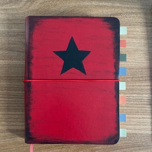 May include: Red journal with a black star on the cover. The journal has a distressed look with a red elastic closure and colorful page tabs. The edges of the cover are dark, giving it a vintage appearance.