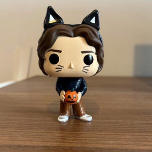 May include: A vinyl collectible figure featuring a character with brown hair, black cat ears, and painted whiskers on the face. The figure wears a dark blue long-sleeved shirt and brown pants, holding a small orange jack-o'-lantern pumpkin in its hands.