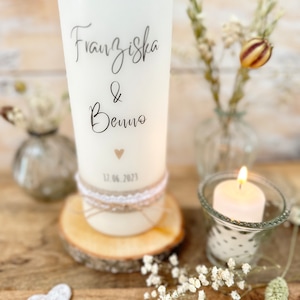 May include: White pillar candle with the names Franziska & Benno and the date 12.06.2023 written on it. The candle is decorated with a small heart and a rustic twine and lace band. The candle is sitting on a wooden slice.