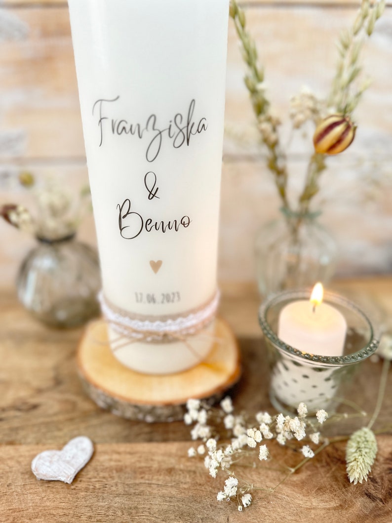 May include: White pillar candle with the names Franziska & Benno and the date 12.06.2023 written on it. The candle is decorated with a small heart and a rustic twine and lace band. The candle is sitting on a wooden slice.