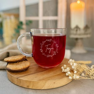 Op de afbeelding: Een heldere glazen mok gevuld met een rode vloeistof, mogelijk thee, met de naam "Tanya" in een bloemenkrans. De mok staat op een houten serveerplank met koekjes. Een brandende kaars en gedroogde bloemen zijn op de achtergrond.