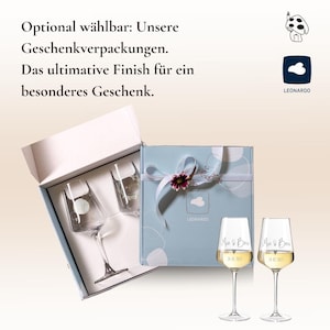 May include: Two clear wine glasses with the text Mia & Ben and a date printed on the bowls, standing next to a light blue gift box containing two additional glasses. The box features a white ribbon, a small flower, and the Leonardo brand logo.
