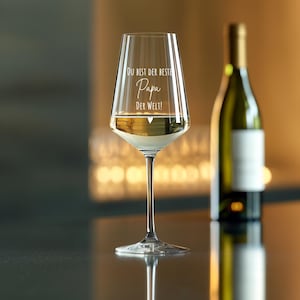 May include: A clear glass wine glass with a white inscription that reads "Du bist der beste Papa der Welt!"  The glass is filled with white wine and a small heart is etched into the glass.