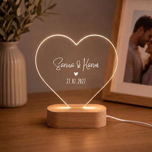 Könnte beinhalten: Ein herzförmiges Acryl-Nachtlicht mit Holzsockel. Das klare Herz zeigt den weiß gravierten Text "Sarina & Karim" über einem kleinen Herzsymbol und dem Datum "27.07.2027". Die Kanten des Herzens leuchten in einem warmen Licht.