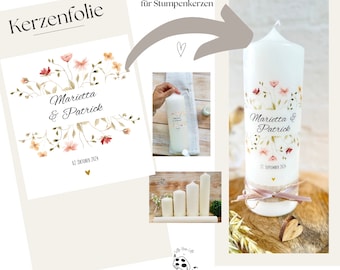 May include: A white candle with a floral design and the text "Marietta & Patrick" and "21. SEPTEMBER 2024". The candle is on a wooden surface with a heart-shaped piece of wood.