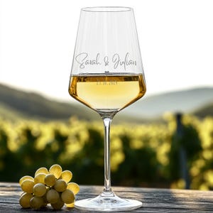 May include: A clear glass wine glass filled with white wine, featuring the engraved names Sarah & Julian and the date 23.10.2024 with a small heart symbol between the names and the date. The glass has a long stem and a wide base.