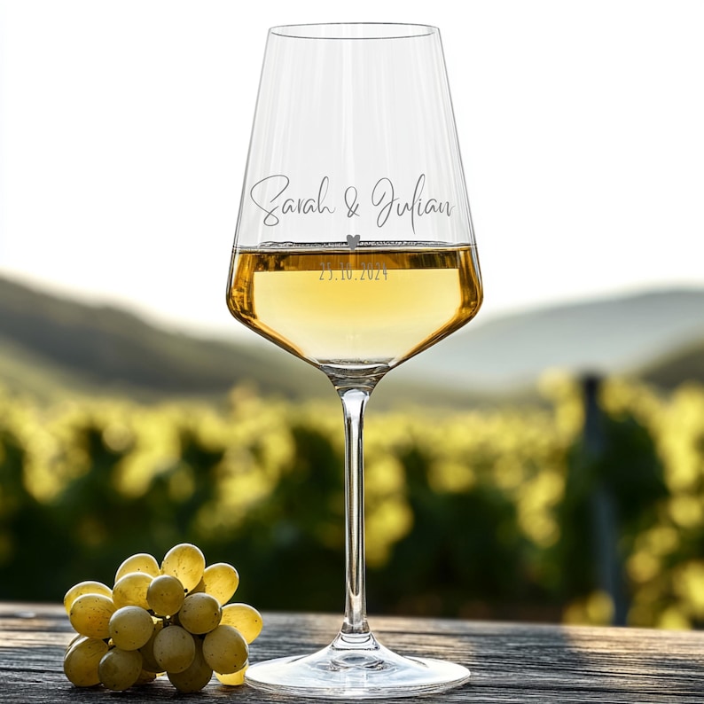 May include: A clear glass wine glass filled with white wine, featuring the engraved names Sarah & Julian and the date 23.10.2024 with a small heart symbol between the names and the date. The glass has a long stem and a wide base.