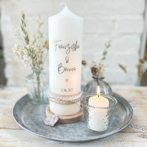 May include: White pillar candle with the names Franziska & Benno and the date 17.06.2023 written on it. The candle is decorated with a small heart and a lace ribbon. It is sitting on a wooden base and a small heart-shaped decoration.