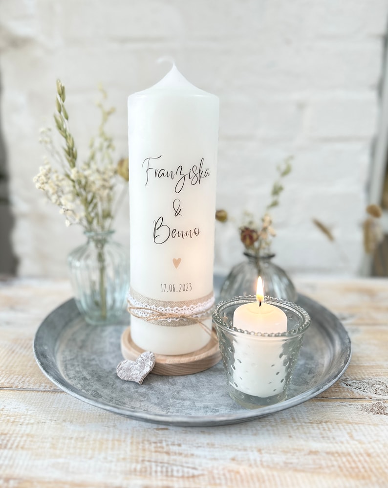 May include: White pillar candle with the names Franziska & Benno and the date 17.06.2023 written on it. The candle is decorated with a small heart and a lace ribbon. It is sitting on a wooden base and a small heart-shaped decoration.