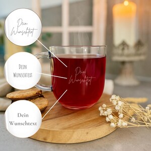 Könnte beinhalten: Eine transparente Glas-Tasse gefüllt mit einer roten Flüssigkeit, möglicherweise Tee oder Saft, mit dem Text "Dein Wunschtext" in einer Schriftart. Die Tasse steht auf einem Holzbrett mit Keksen und Trockenblumen. Eine brennende Kerze ist im Hintergrund.