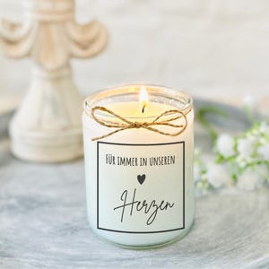 Op de afbeelding: Een witte kaars met een bruine touw strik en een label met de tekst "Für immer in unseren Herzen" met een klein hart symbool.