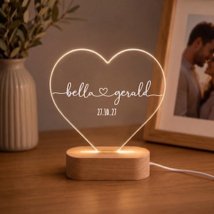 Può includere: Una lampada in acrilico a forma di cuore con base in legno. L'acrilico trasparente presenta il testo inciso in bianco "bella" e "gerald" separati da una piccola icona a forma di cuore, con la data "27.10.27" sotto. La luce emette un bagliore caldo.