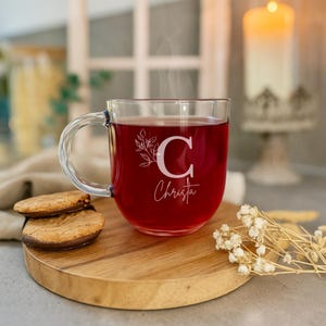 Op de afbeelding: Een heldere glazen mok gevuld met een rode vloeistof, mogelijk thee of sap, met stoom die opstijgt. De mok heeft een handvat en is versierd met een bloemenontwerp en de letter "C" en de naam "Christa". Twee koekjes liggen op een houten plank.