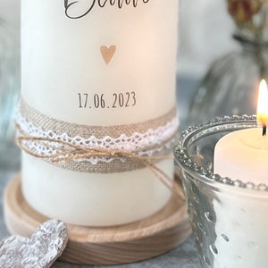 May include: White candle with a burlap and lace ribbon, a small heart, and the date 17.06.2023 printed on the side. The candle is on a wooden base and next to a glass candle holder with a lit candle inside.