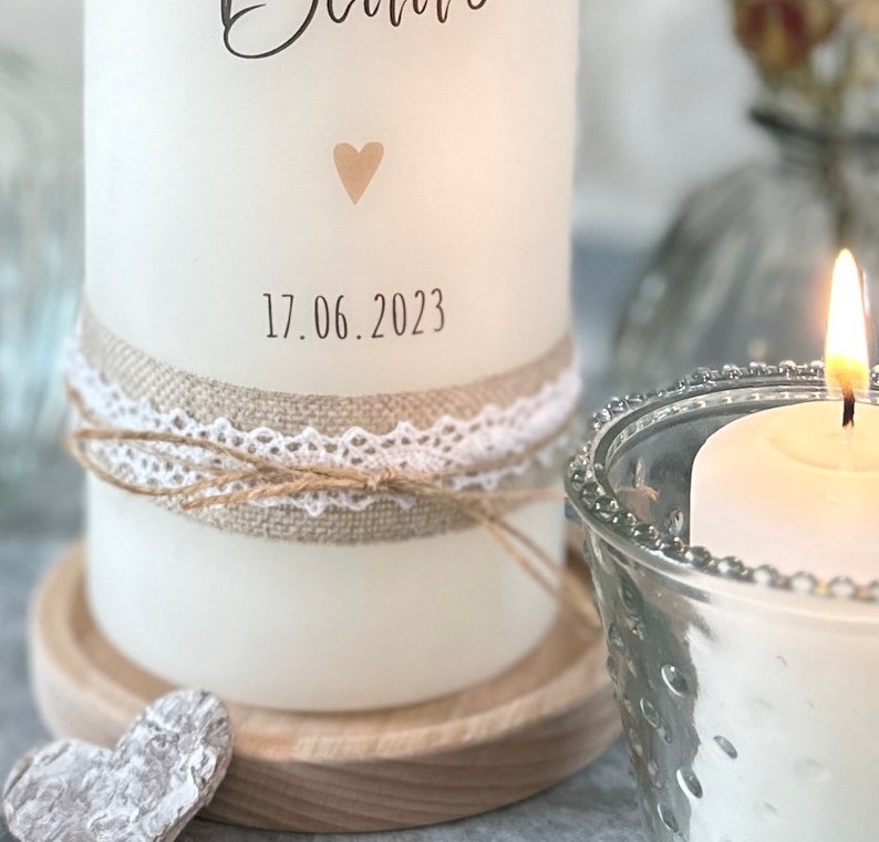 May include: White candle with a burlap and lace ribbon, a small heart, and the date 17.06.2023 printed on the side. The candle is on a wooden base and next to a glass candle holder with a lit candle inside.