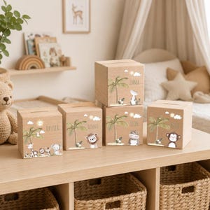 Könnte beinhalten: Fünf quadratische Spardosen aus hellem Holz mit Münzschlitzen auf der Oberseite. Jede Box zeigt eine aufgedruckte Illustration mit einer Palme, einer Sonne, Wolken und einem anderen Tier, darunter eine Giraffe, ein Löwe, ein Nilpferd und ein Affe.