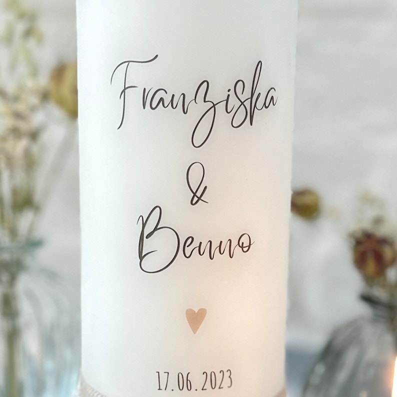 May include: White candle with the names Franziska & Benno and the date 17.06.2023 printed on it. A small heart is printed below the names.