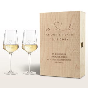 Puede incluir: Dos copas de vino transparentes llenas de un líquido claro, junto a una caja de madera. Las copas y la caja están grabadas con los nombres "ANOUK & HEATH" y la fecha "12.11.2024". La caja tiene un diseño de corazón.