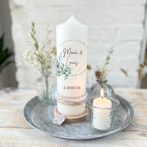 Könnte beinhalten: Eine weiße Säulenkerze mit goldenem und grünem Blumendesign, den Namen "Mona & Louis" und dem Datum "02. Oktober 2026". Die Kerze ist mit Spitze und Jute verziert und steht auf einem Holzsockel. Eine brennende Votivkerze befindet sich in einem Glas.