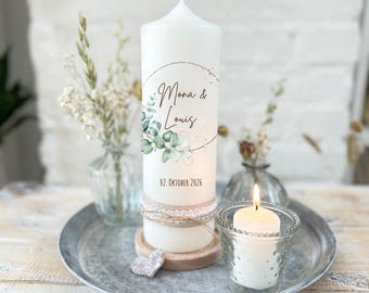 Peut inclure: Une bougie pilier blanche avec un motif floral doré et vert, les noms "Mona & Louis" et la date "02. Oktober 2026". La bougie est décorée de dentelle et de toile de jute et repose sur une base en bois. Une bougie chauffe-plat allumée est dans un verre.