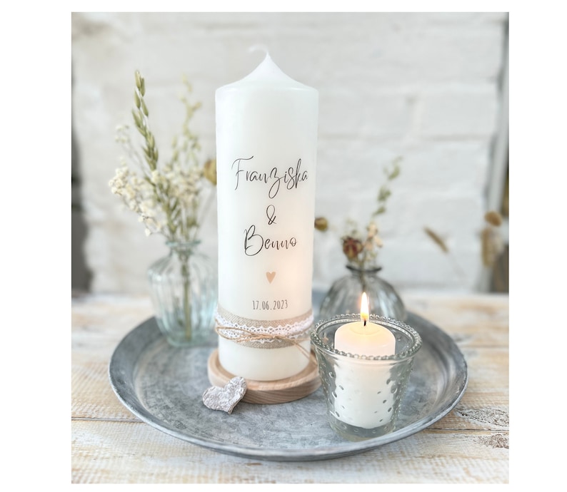 May include: White pillar candle with a decorative lace trim and the text "Franziska & Benno 17.06.2023" in black script. The candle is on a wooden base and there is a small heart-shaped decoration on the candle.